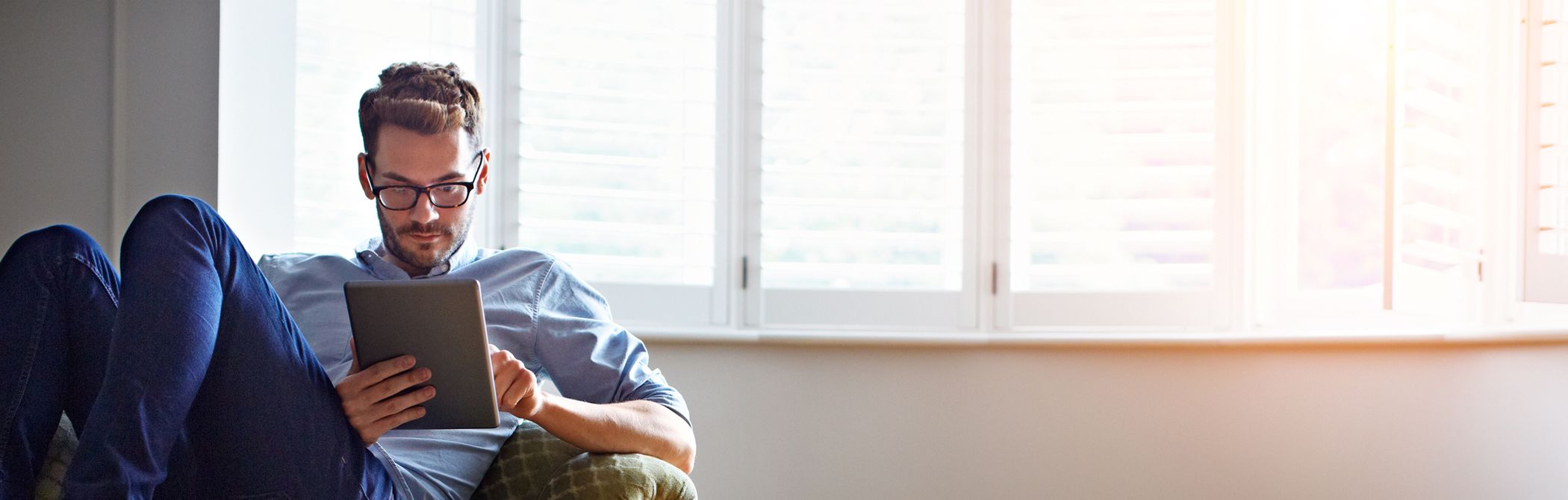 A Millennial reads his tablet in front of some windows.